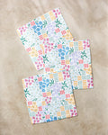 Three "Floral Garden" paper napkins by Charlie Rowan Designs, featuring colorful floral and geometric patterns on recycled paper, are arranged in a staggered layout on a beige marble surface.