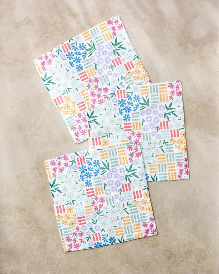 Three "Floral Garden" paper napkins by Charlie Rowan Designs, featuring colorful floral and geometric patterns on recycled paper, are arranged in a staggered layout on a beige marble surface.