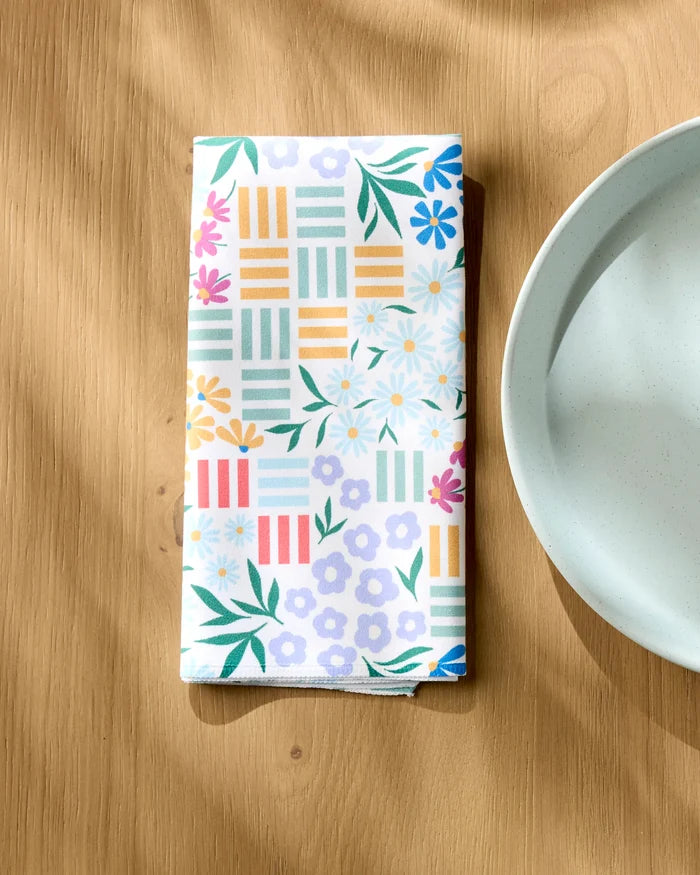 A folded Charlie Rowan Designs "Floral Garden" eco-friendly napkin with vibrant floral and geometric patterns rests on a wooden table by a white plate, sunlight highlighting the sustainable choice of this Dinner Napkin Set.