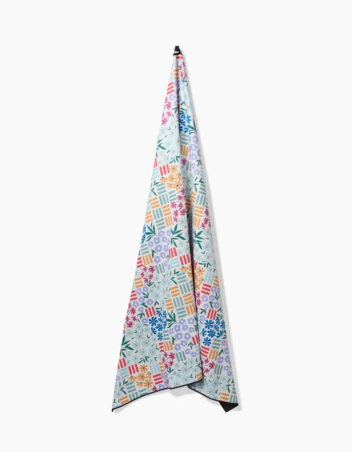 The Floral Garden quick-drying towel by Charlie Rowan Designs, featuring a vibrant geometric and floral pattern, hangs vertically on a white wall with its pointed end facing up.