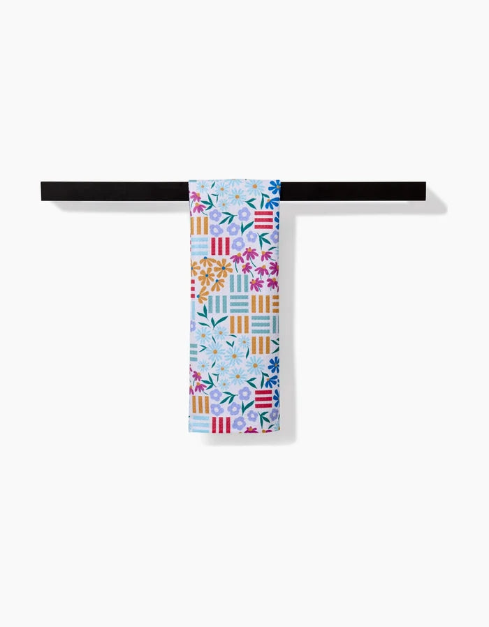 The Floral Garden bar towel by Charlie Rowan Designs features vibrant floral and geometric patterns in blue, orange, pink, and yellow on soft Geoweave fabric, bringing lively color to your space when hung on a black towel bar.