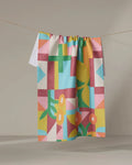 The "Floral Geometry" recycled kitchen towel by Lisa Nelson hangs from a string against a neutral background, showcasing vibrant geometric and floral patterns in pink, blue, yellow, green, and orange.