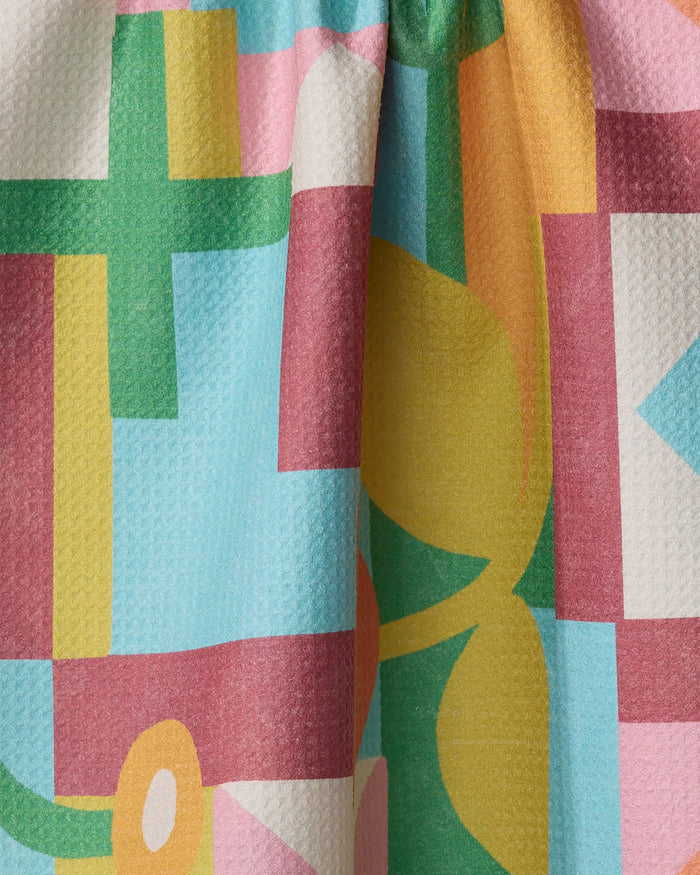 A close-up of Lisa Nelson's "Floral Geometry" tea towel shows its colorful geometric pattern in green, yellow, pink, and blue, softly draped with gentle folds on a textured surface.