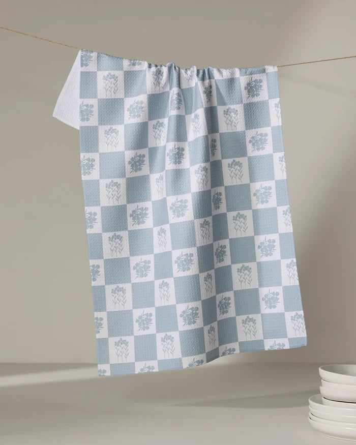 The Charlie Rowan Designs Floral Patchwork tea towel, featuring blue and white checks with floral patterns and made from recycled materials, hangs indoors near stacked white plates on a light surface.