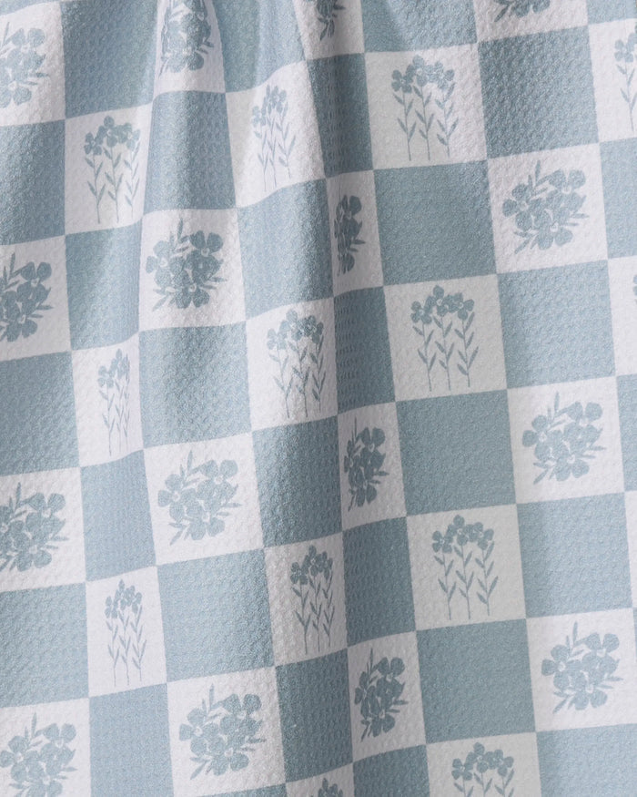 The Floral Patchwork tea towel by Charlie Rowan Designs features a blue and white checkered pattern with floral accents. Draped folds reveal its textured surface, and it's crafted from recycled materials.