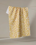 The Gerdadzy "Floral Spell" tea towel, featuring a yellow and white floral pattern and made from recycled materials, hangs on a clothesline against a plain neutral background.