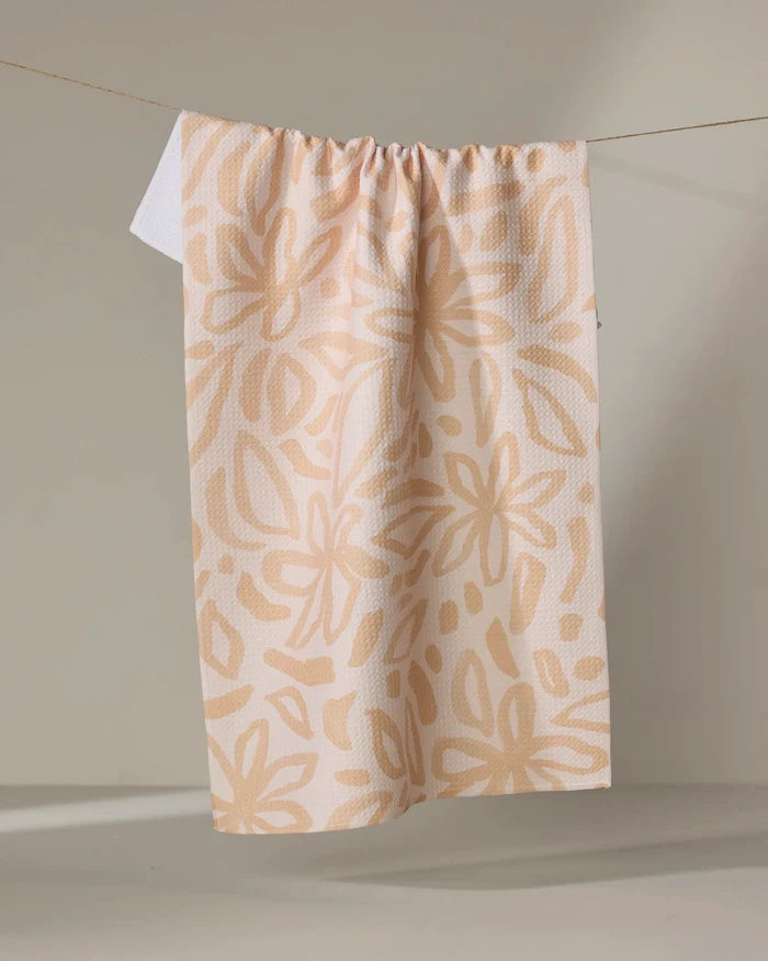 The Jen B. Peters Floral towel, an eco-friendly beige kitchen towel with a large abstract light tan floral pattern, hangs from a string against a neutral background.