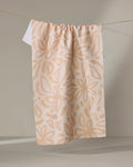 The Jen B. Peters Floral towel, an eco-friendly beige kitchen towel with a large abstract light tan floral pattern, hangs from a string against a neutral background.
