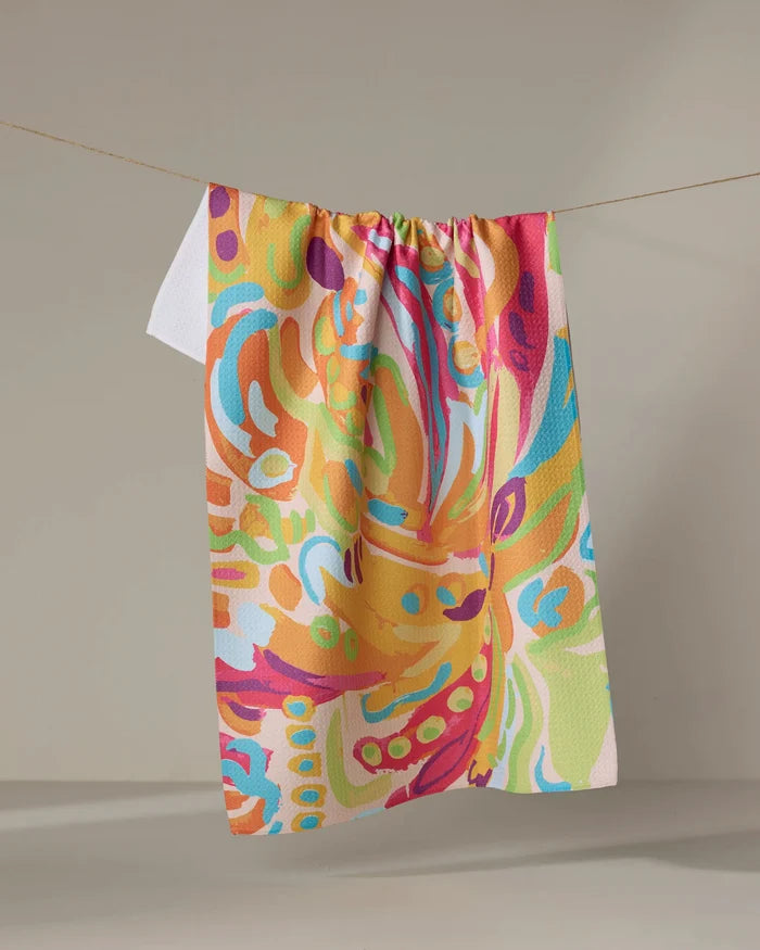 The Florescence tea towel by Sareka Unique features an abstract, vibrant pattern in orange, pink, yellow, green, blue, and white. Made from quick-drying recycled materials, it hangs on a clothesline against a neutral background.