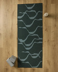 A GEOMETRY Flow yoga mat in dark green with light gray wavy lines lies on a wooden floor. Nearby are a recycled yoga towel, two gray yoga blocks in the bottom left, and a beige water bottle at the top right of the mat.