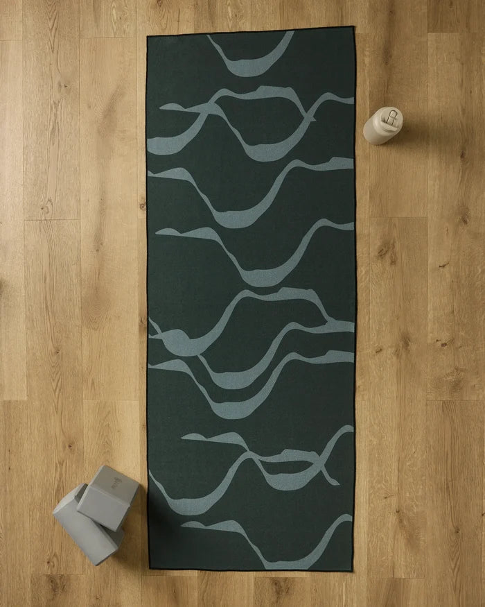 A GEOMETRY Flow yoga mat in dark green with light gray wavy lines lies on a wooden floor. Nearby are a recycled yoga towel, two gray yoga blocks in the bottom left, and a beige water bottle at the top right of the mat.