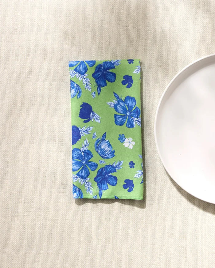 The GEOMETRY Flower Bomb reusable napkin, featuring a blue and white floral pattern on green fabric, is neatly folded on a beige textured surface beside a partially visible white plate.