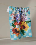 The Flower Bouquet tea towel by Pascale Dilger features a blue and white checkered pattern with vibrant sunflowers and colorful flowers, displayed hanging on a clothesline against a neutral background. Ultra-absorbent and stylish.