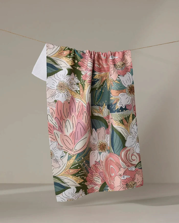 The Flower Explosion tea towel from Second Nature Studio features a vibrant floral print in pink, green, white, and yellow. Made from ultra-absorbent fabric for everyday use, it hangs on a clothesline against a plain beige background.