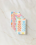 Five reusable Flower Parade paper towels by Lizzie Clark, featuring pastel floral and leaf designs in blue, pink, and orange, are stacked on a white marble surface with light brown veining.
