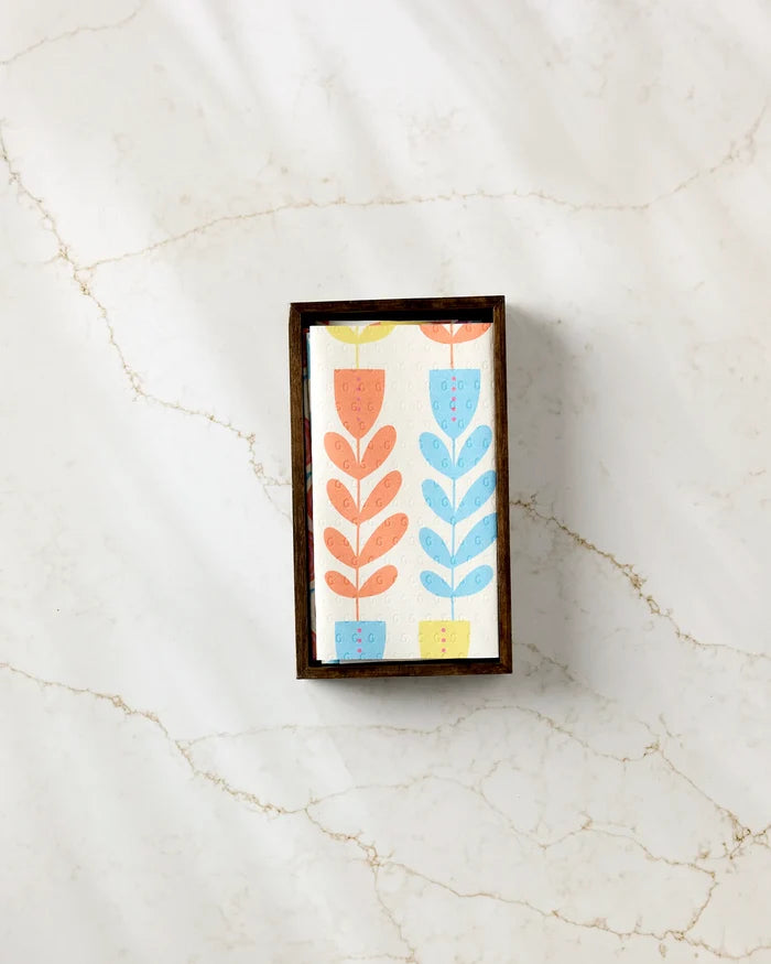 The Flower Parade soap bar by Lizzie Clark, featuring stylized orange, yellow, and blue flowers, rests in a dark wooden dish on white marble with tan veins, next to a stack of eco-friendly paper towels.