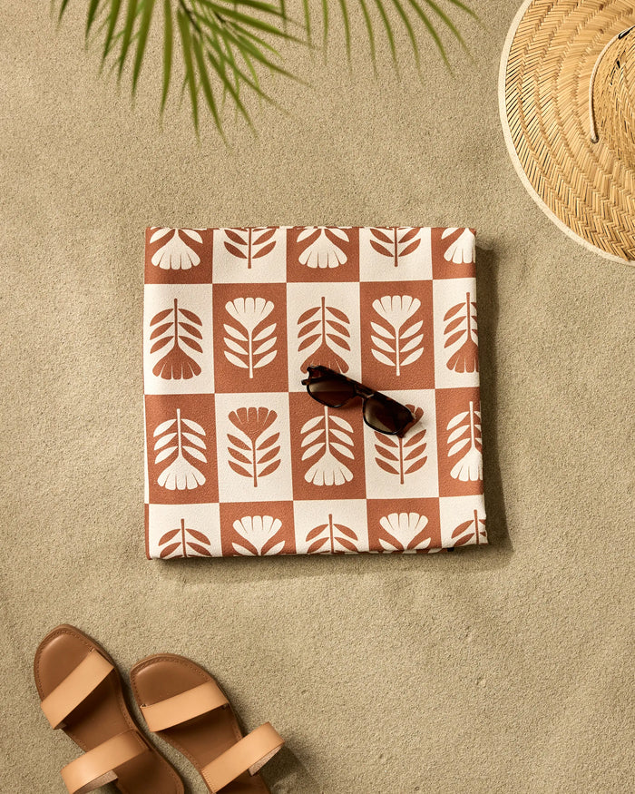 The Flower Patch quick-drying beach blanket from Charlie Rowan Designs, folded with a floral print and sunglasses on top, rests on the sand beside tan sandals, a woven hat, and a palm leaf casting a shadow.