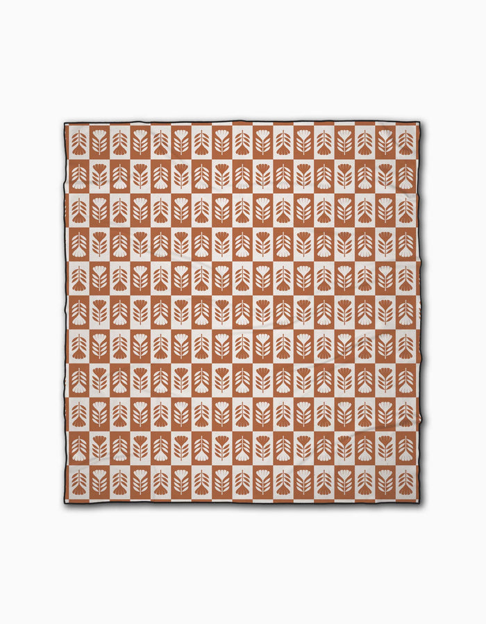 The Flower Patch Beach Blanket by Charlie Rowan Designs is a square micro-loop blanket displaying orange squares with white stylized leaf patterns, arranged in a grid on a white background.