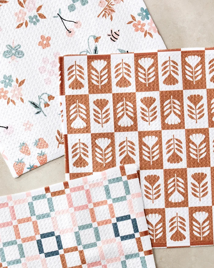 Three Flower Patch dishcloths by Charlie Rowan Designs, made from recycled materials, are shown on a light surface: one with fruit, flowers, and bees; one with a brown geometric floral pattern; and one with pastel pink, blue, and brown squares.