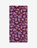 The Flower Pop napkin by Elisabeth Wing is a rectangular cloth made from recycled materials, featuring a dark purple background with light purple daisies and orange centers scattered across the surface.