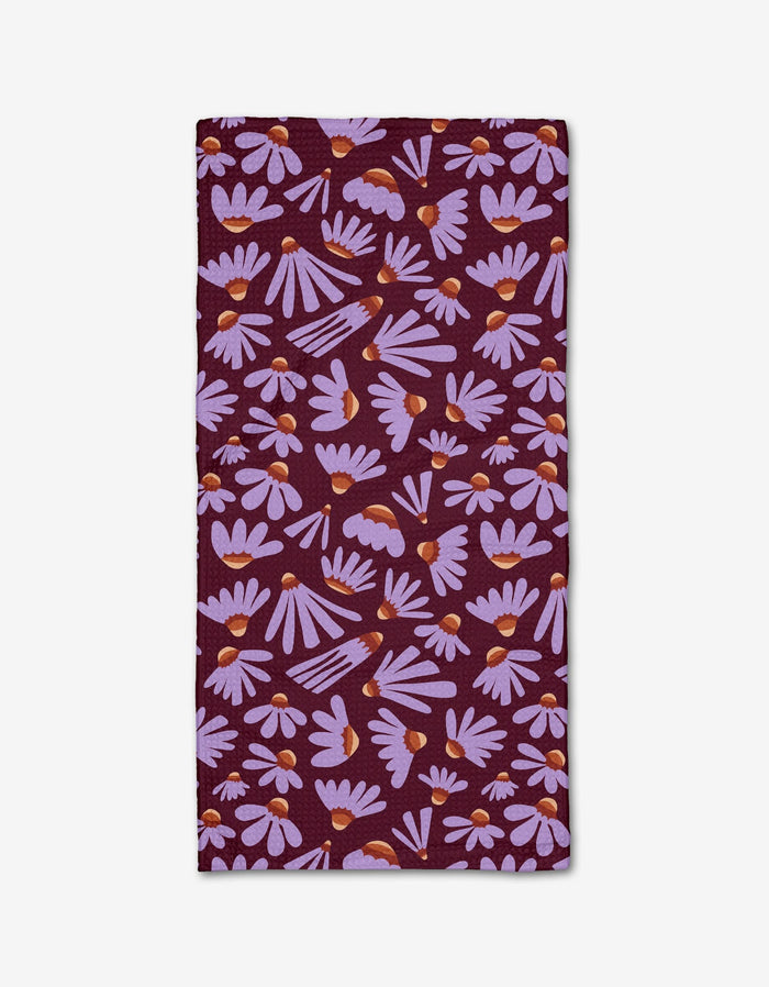 The Flower Pop napkin by Elisabeth Wing is a rectangular cloth made from recycled materials, featuring a dark purple background with light purple daisies and orange centers scattered across the surface.