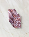 Six Flower Pop cloth napkins by Elisabeth Wing, featuring a light purple and white floral pattern on mauve, are stacked diagonally on marble—a stylish, eco-friendly alternative to kitchen and paper towels.