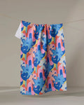 The Flower Powers towel by Masha Volnova, featuring a playful blue and pink floral abstract pattern, hangs on a clothesline against a neutral background.