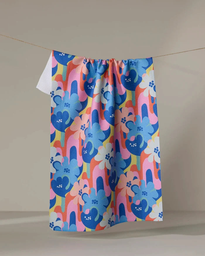 The Flower Powers towel by Masha Volnova, featuring a playful blue and pink floral abstract pattern, hangs on a clothesline against a neutral background.