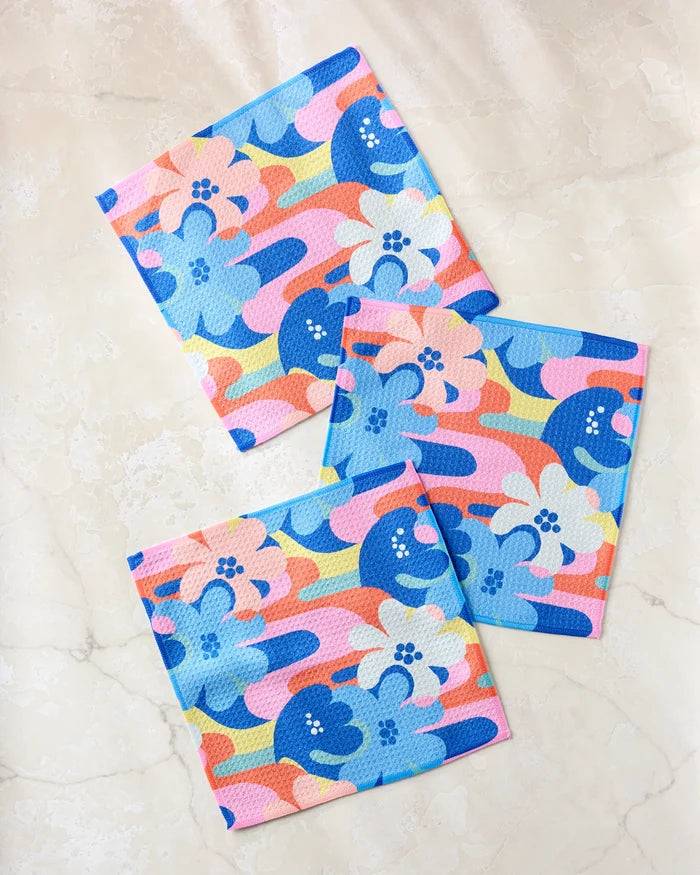 The Flower Powers dishcloth set by Masha Volnova includes three super-absorbent towels with abstract floral patterns in blue, pink, yellow, and orange, beautifully displayed on a light beige marble surface.