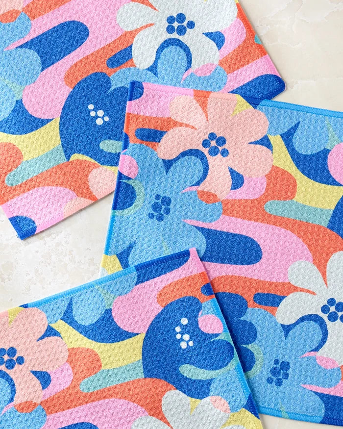 Three "Flower Powers" cloths by Masha Volnova, crafted from recycled materials with abstract floral patterns in blue, pink, orange, yellow, and white, overlap on a light marble surface.