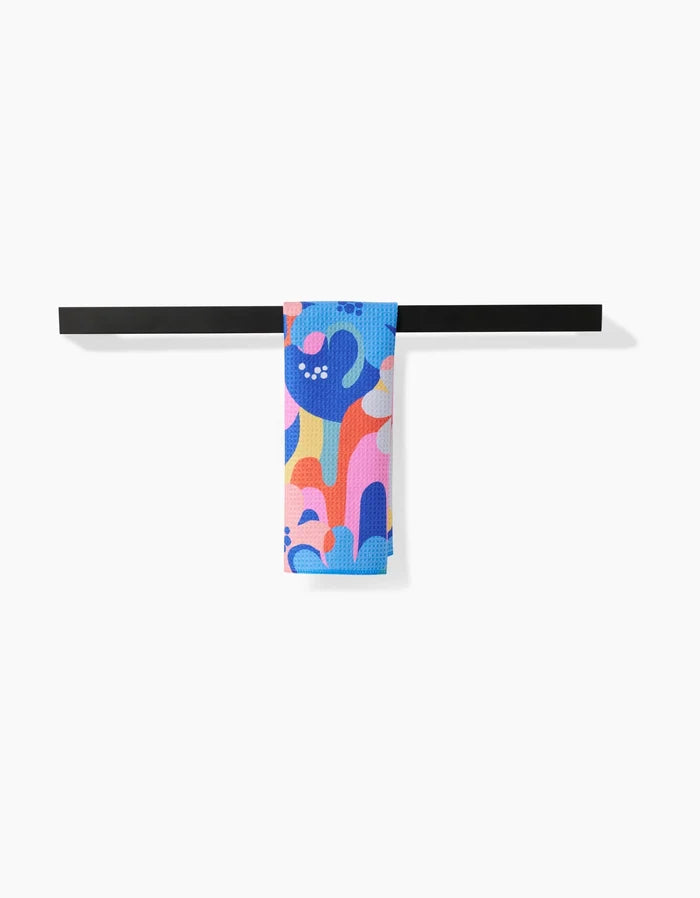 The Flower Powers Bar Towel by Masha Volnova features abstract blue, pink, orange, and purple shapes on soft Geoweave fabric. It hangs neatly over a black rod against a white background, offering superior absorbency.