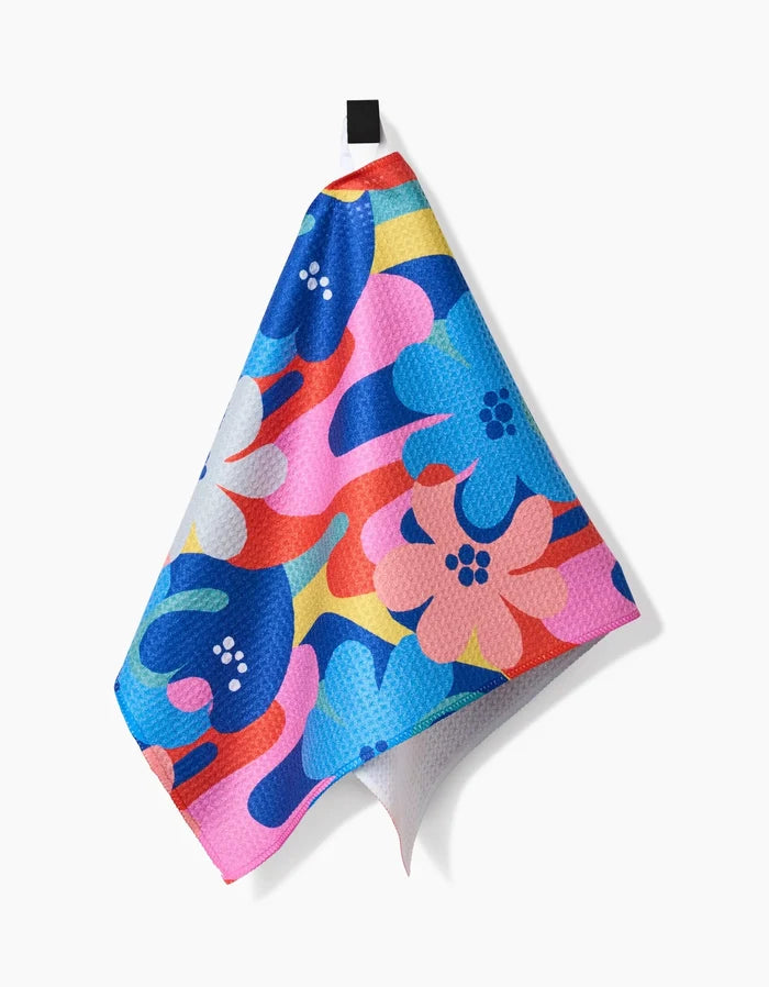 The Flower Powers bar towel by Masha Volnova displays a bold, abstract floral design in blue, pink, red, white, and yellow. Made from recycled materials, it hangs from a black loop and adds vibrant style to any setting.