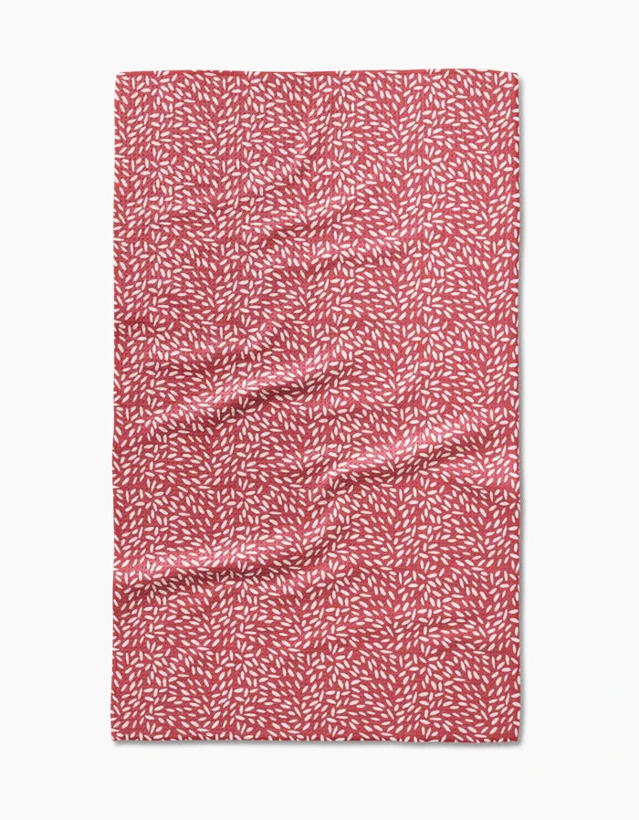 A Flower Seeds tea towel by Amita Nair, made of Geoweave fabric in red with a pattern of small white leaf-like shapes, shown laid flat with slight surface wrinkles.