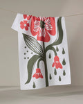 The "Flower Stamp" tea towel by Ash Cascade, made from recycled materials, features bold red flower graphics with green leaves and droplets, hanging on a clothesline against a neutral backdrop.