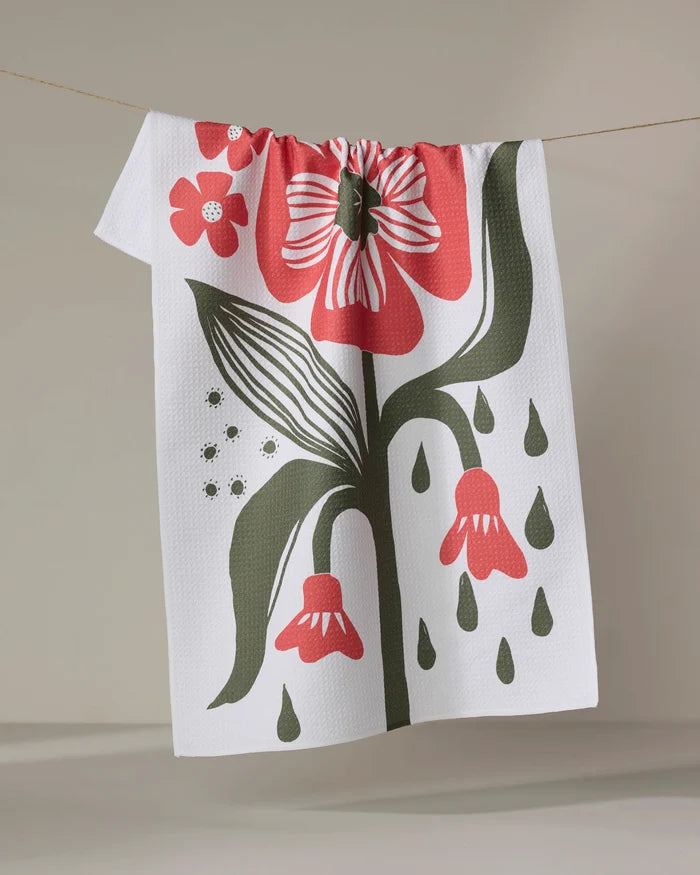 The "Flower Stamp" tea towel by Ash Cascade, made from recycled materials, features bold red flower graphics with green leaves and droplets, hanging on a clothesline against a neutral backdrop.