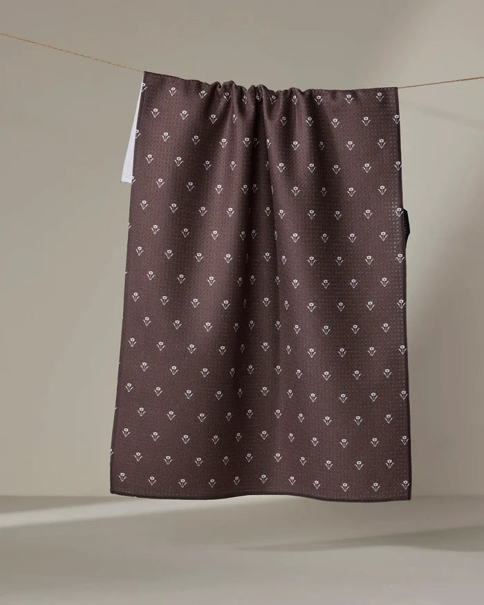 The Buy Guide’s Flowerfield Chocolate tea towel, crafted from recycled materials and featuring white diamond and floral patterns, hangs over a clothesline against a neutral background.