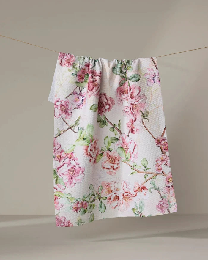 The Pascale Dilger "Flowering Quince" white textured kitchen towel, featuring a pink and green floral print, hangs from a clothesline against a light background.