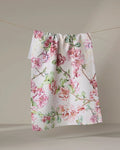 The Pascale Dilger "Flowering Quince" white textured kitchen towel, featuring a pink and green floral print, hangs from a clothesline against a light background.