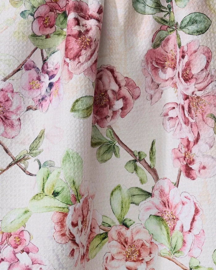 A close-up of the Flowering Quince kitchen towel by Pascale Dilger shows its textured surface with pink and red floral patterns and green leaves on a light background.