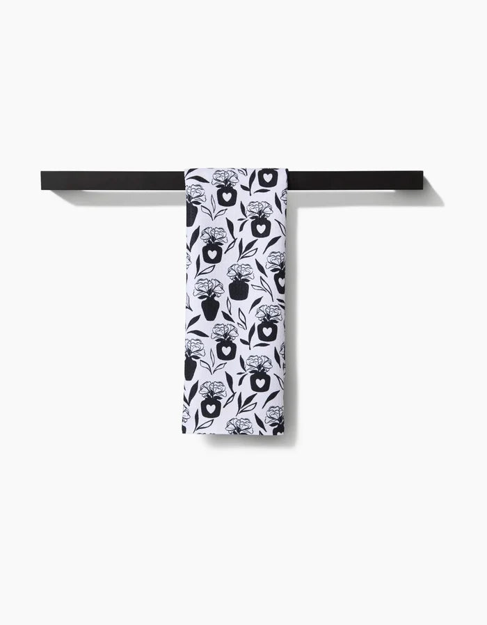 The Flowers for Me tea towel by Charlie Rowan Designs, featuring a black and white floral pattern, is neatly draped over a black towel bar mounted on a white wall.