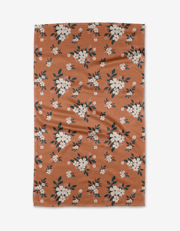 The Flowers in the Field tea towel by Charlie Rowan Designs is a rectangular brown towel with white and pink floral patterns and dark green leaves, crafted from recycled materials and shown flat on a white background.