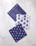 Three Flowing Ocean napkins by Geometry are displayed on white marble: two in navy with white patterns, and one in white with a blue floral design.