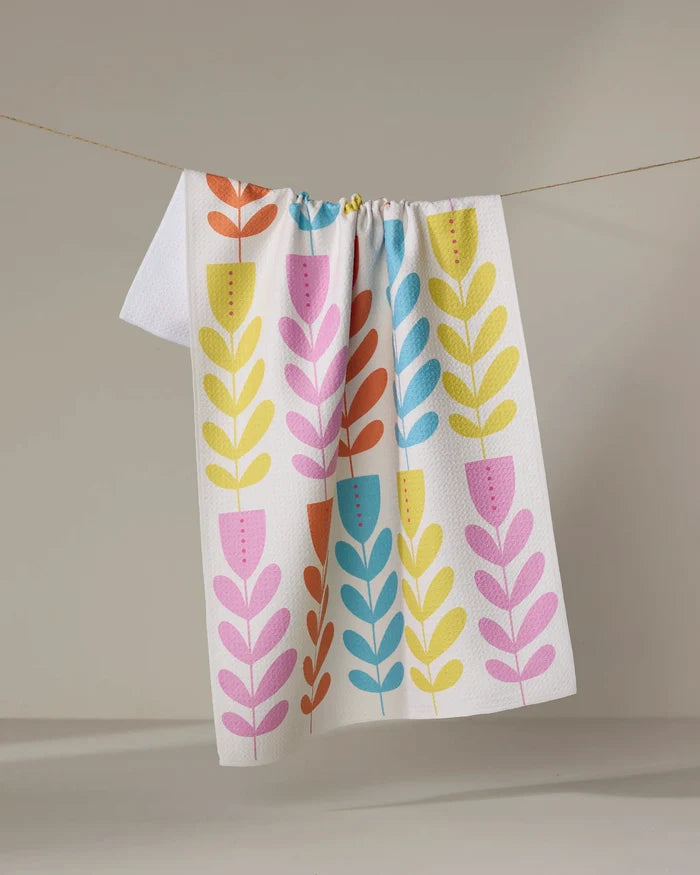 The Folk Art Tulips dish towel by Lizzie Clark, featuring a colorful pattern of stylized leaves and flowers, hangs on a clothesline against a plain beige background.