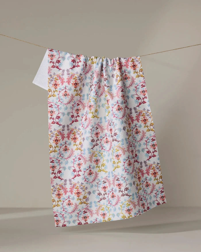 The “Folk Tapestry” kitchen towel by Maayan Alper-Swan, featuring eco-friendly fabric and vibrant red, yellow, and blue floral patterns, hangs on a clothesline against a plain beige background.