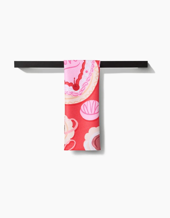 The Food of Love tea towel by Janna Sue Design features bold pink and red abstract florals. Made from ultra-absorbent recycled materials, it’s draped over a black bar against a white background.