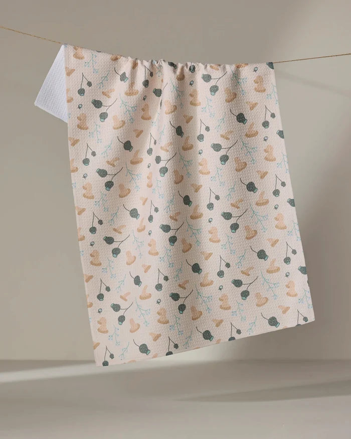 The "Forage" kitchen tea towel by Amberleigh Adoff features a cream color, ultra-absorbent fabric, and a charming pattern of tan rubber ducks with blue and green branches, displayed on a clothesline against a neutral backdrop.