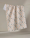 The "Forage" kitchen tea towel by Amberleigh Adoff features a cream color, ultra-absorbent fabric, and a charming pattern of tan rubber ducks with blue and green branches, displayed on a clothesline against a neutral backdrop.