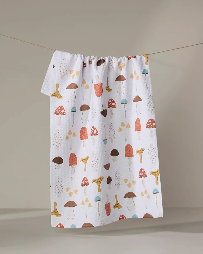 The "Foraging" recycled kitchen towel by Sara Mari, decorated with red, brown, yellow, and blue mushrooms, hangs on a clothesline against a neutral background.
