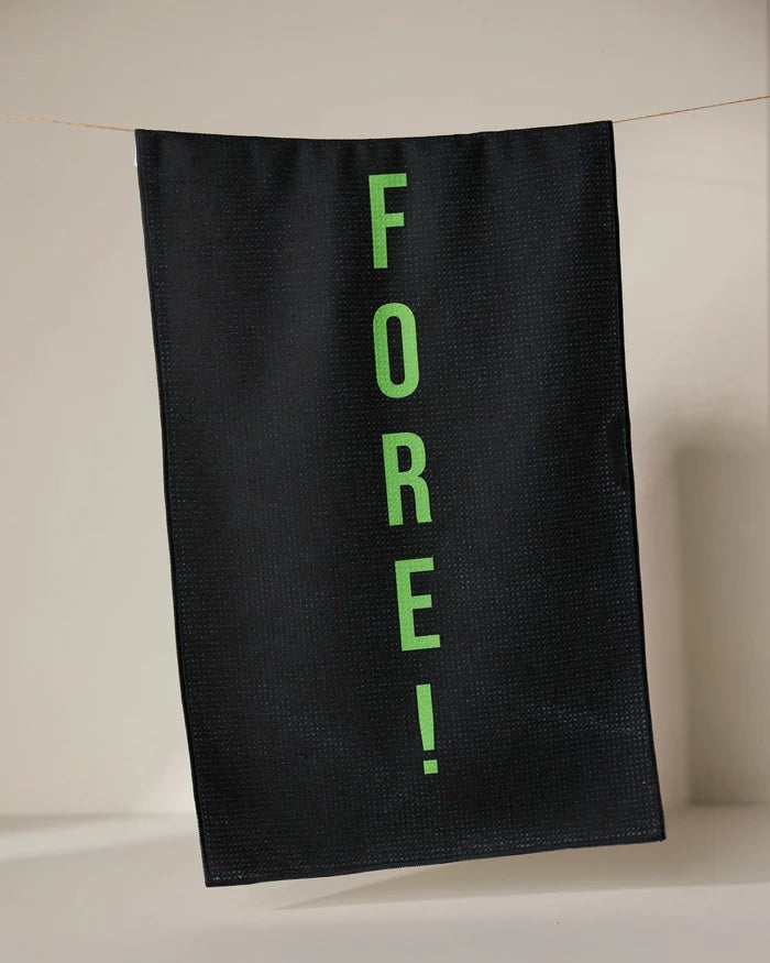 The GEOMETRY Fore tea towel, made from recycled materials, dries quickly and features FORE! in bright green letters printed vertically on black fabric, hanging from a thin line against a neutral background.