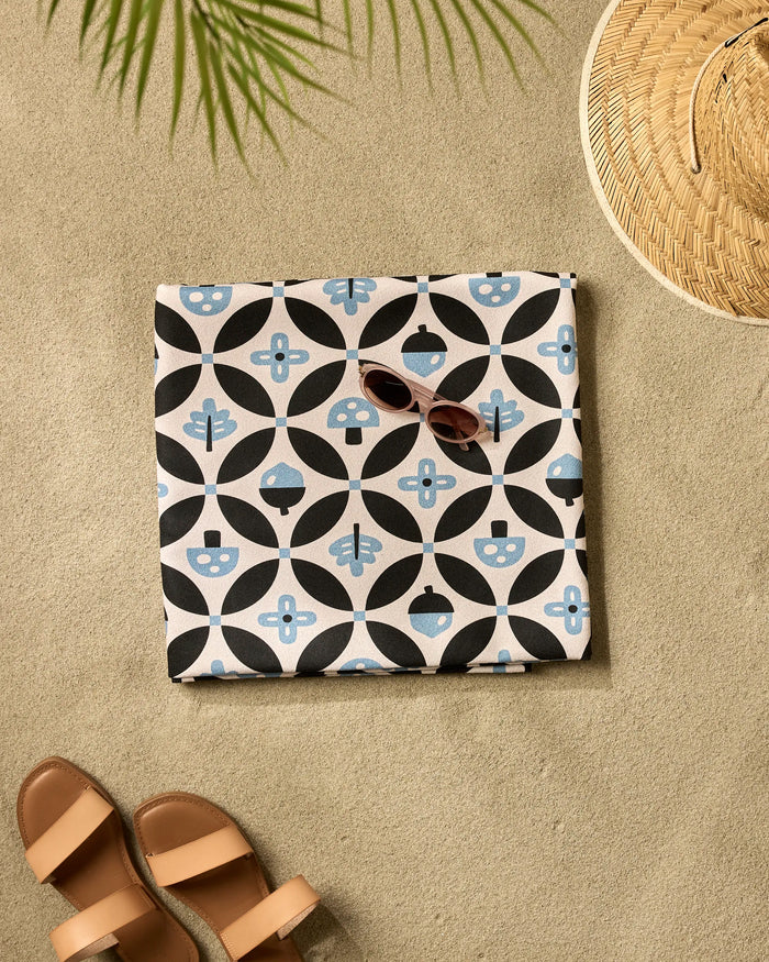 A towel and sunglasses rest on the floor beside the Forest Findings quick-drying beach blanket by Anette Heiberg, perfect for your next seaside adventure.